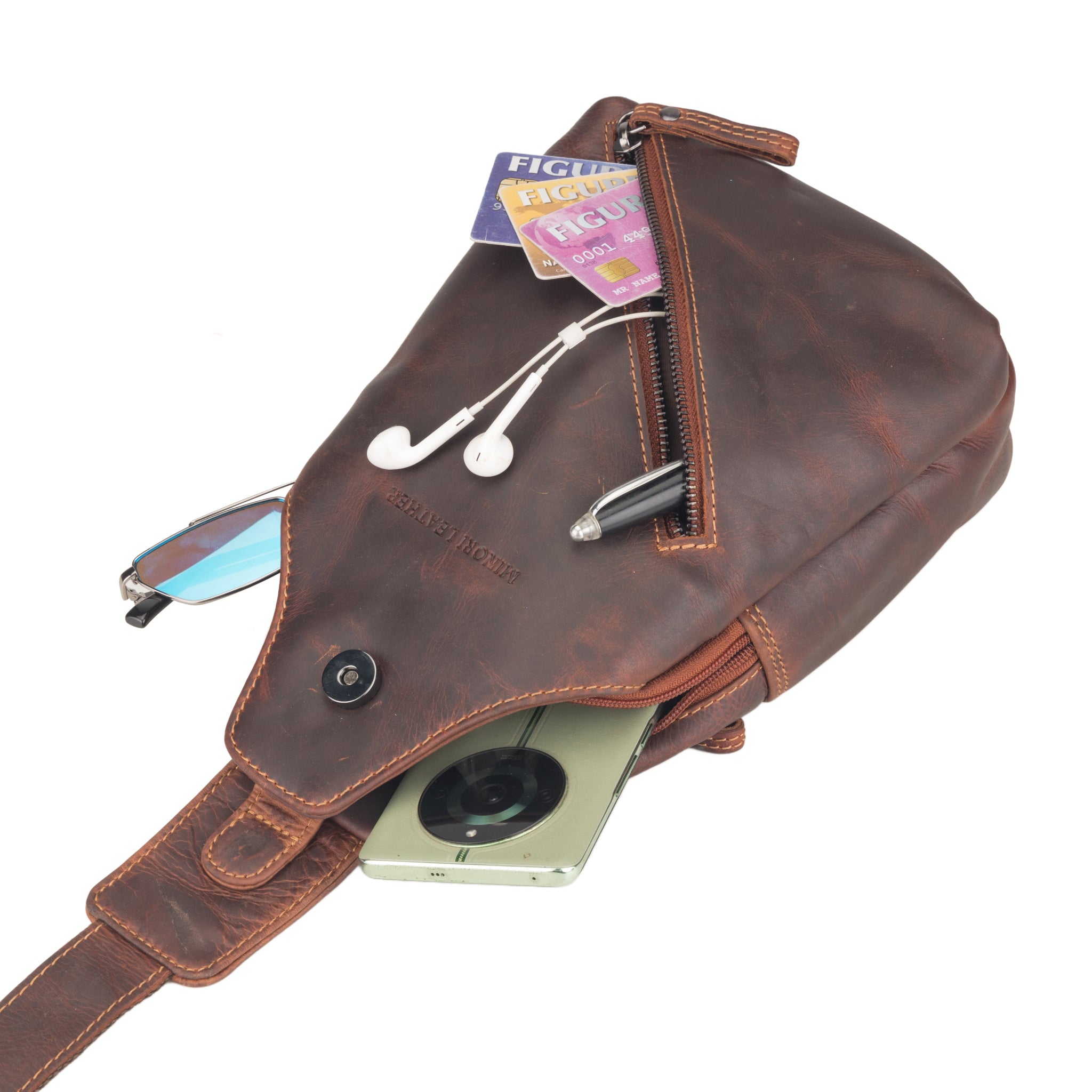 Chestnut brown Minori leather sling bag shown with accessories like phone, earphones, cards, and glasses partially inside compartments.