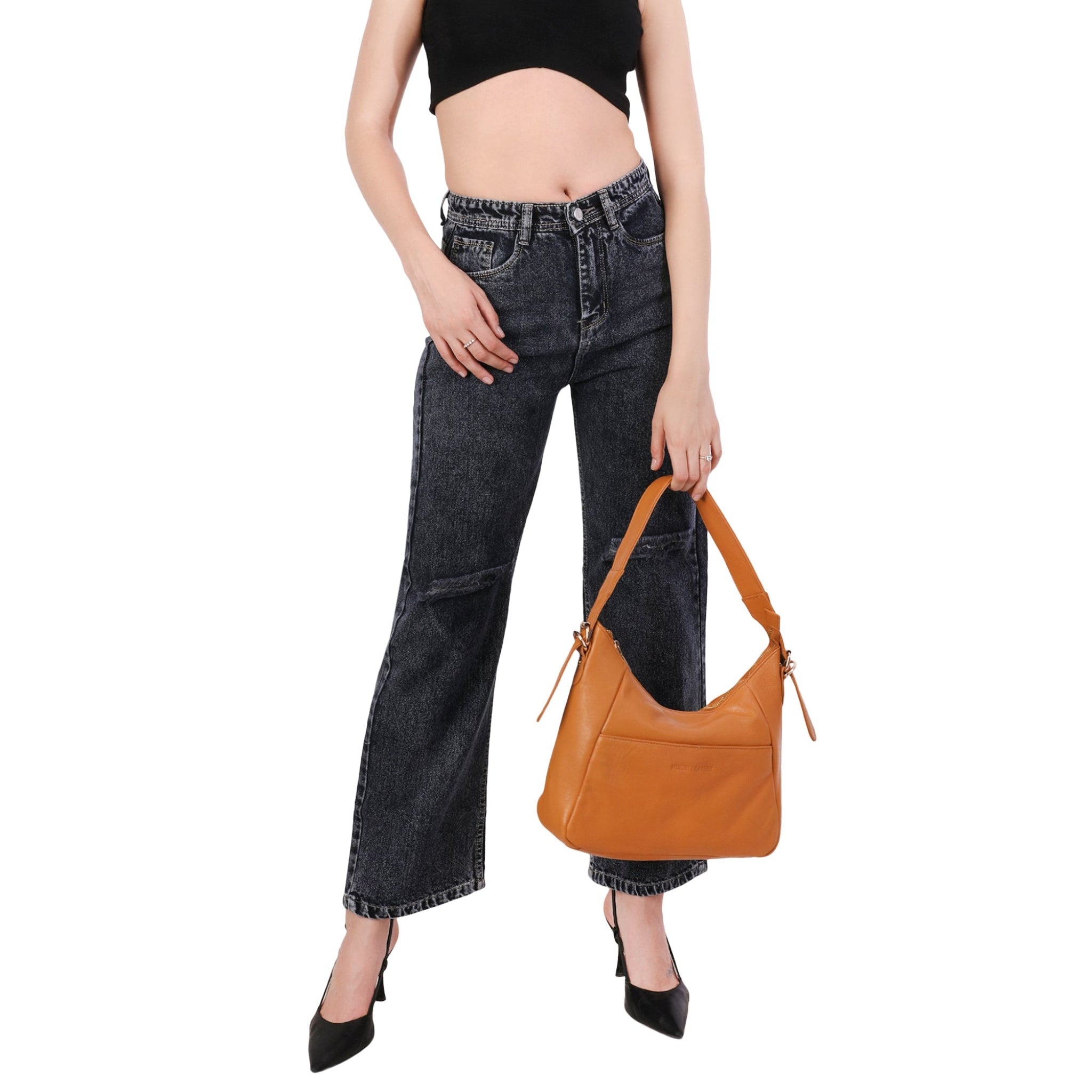 Woman wearing a black crop top and black denim jeans, holding a tan Minori leather shoulder bag with structured design and wide strap.