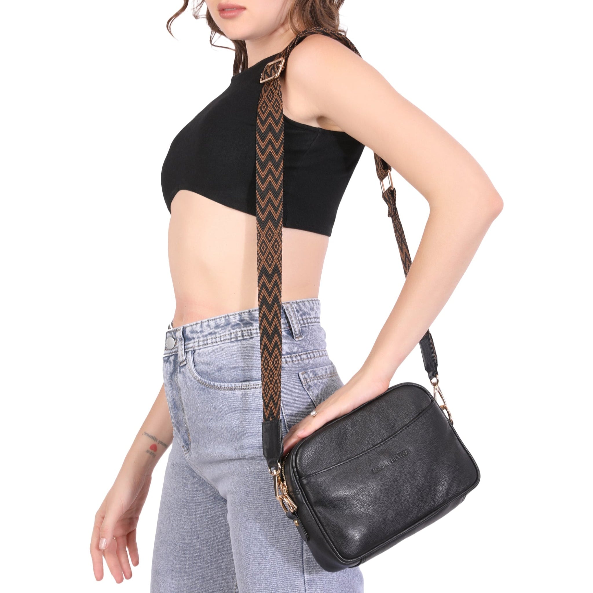 Side profile of a woman wearing a black crop top and jeans, showcasing a black Minori leather crossbody bag with a wide, patterned shoulder strap.