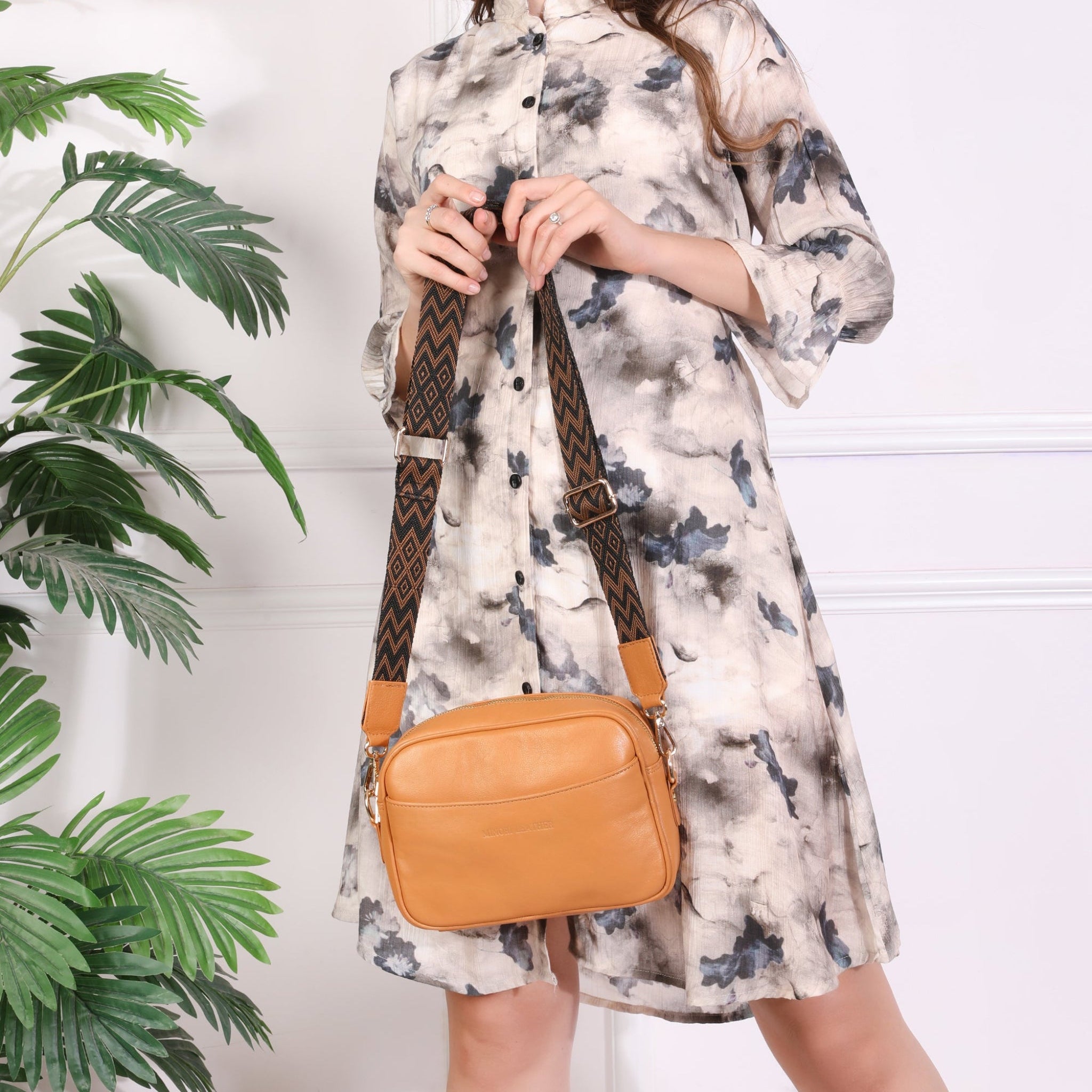 Woman wearing a tie-dye buttoned dress holding a tan Minori leather crossbody bag with a patterned adjustable strap, smiling and standing beside indoor plants.
