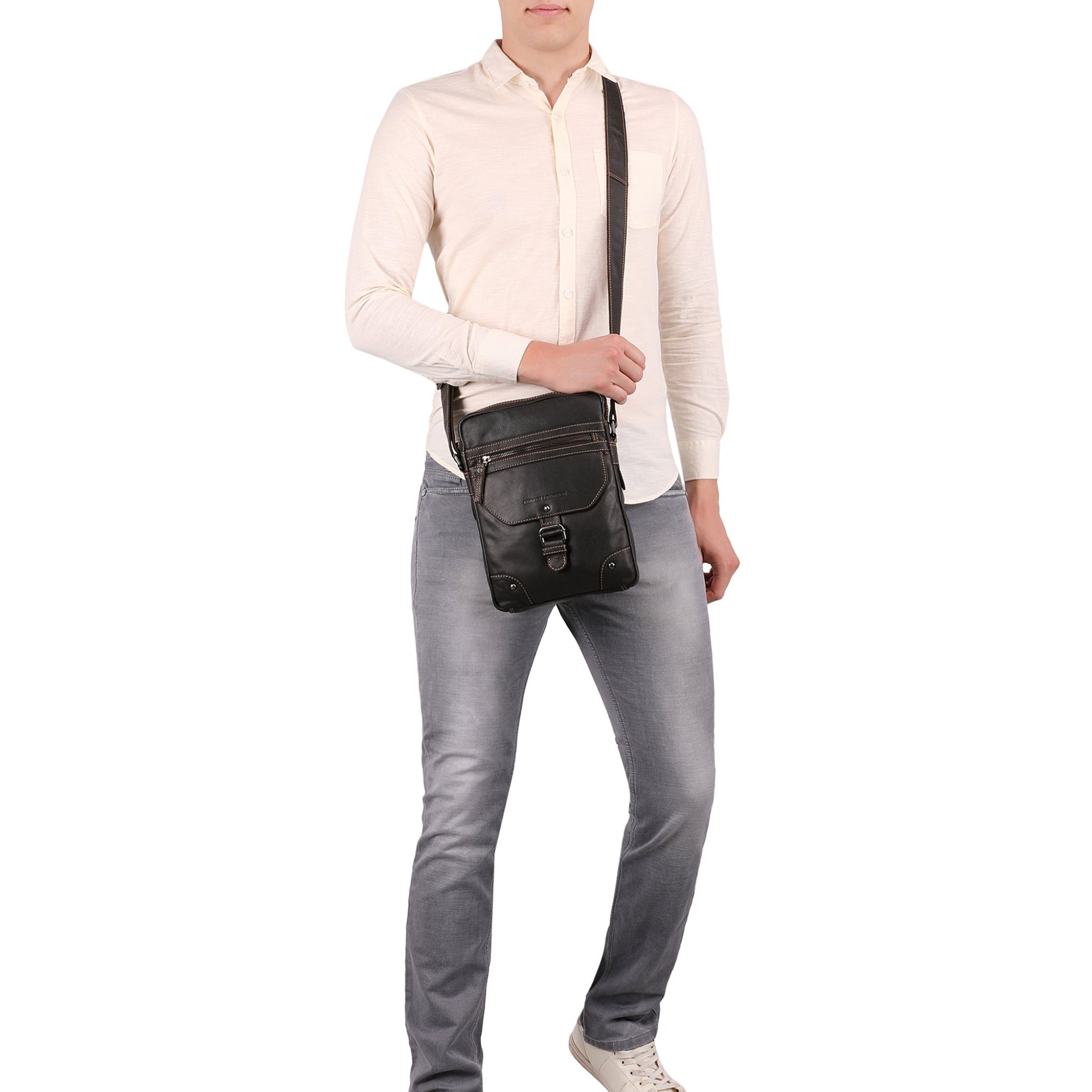 Full-body shot of a man wearing a cream shirt and grey jeans, carrying a dark Minori leather crossbody bag across his chest.