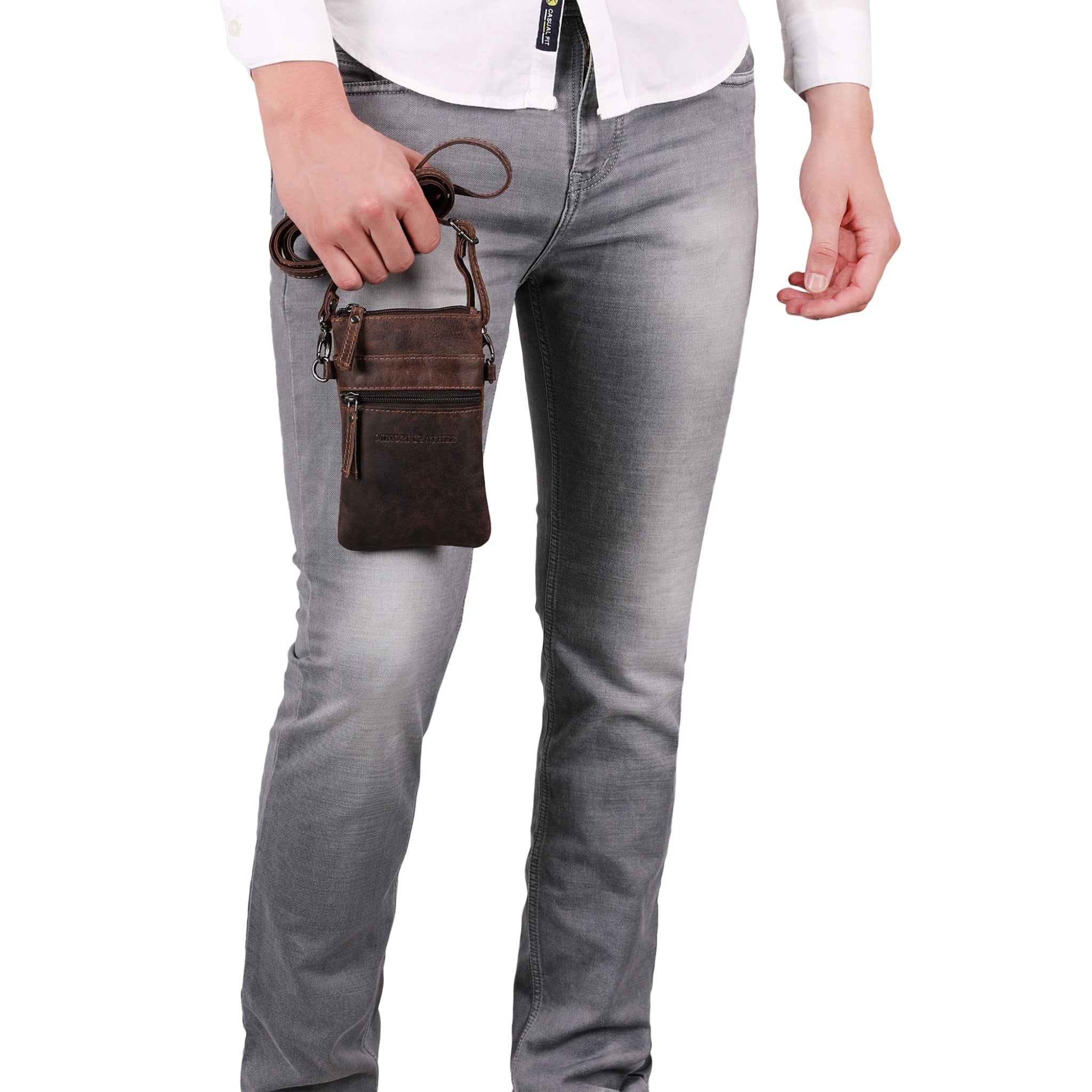 Close-up of man’s lower torso and legs, holding a brown Minori leather crossbody bag by the strap near his hip.