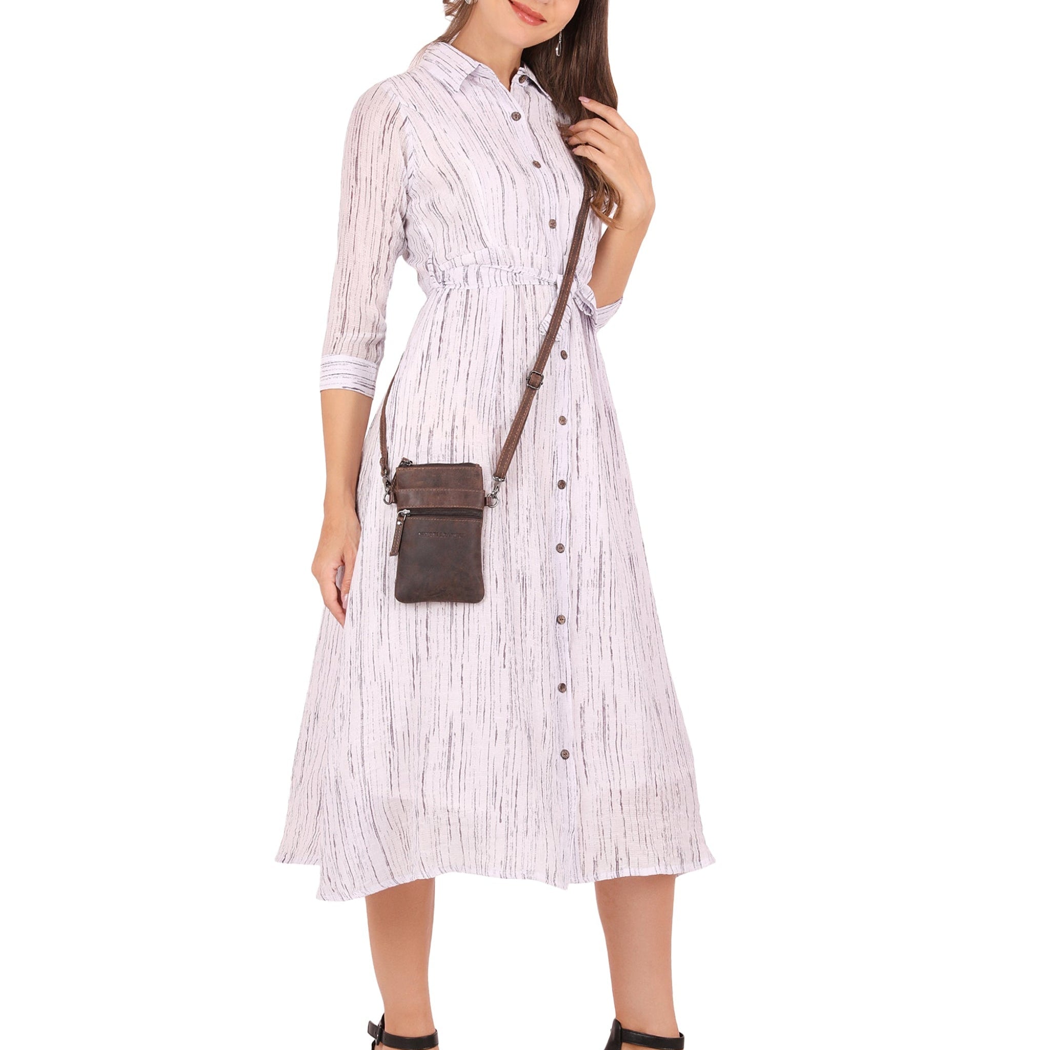 A chestnut brown Minori leather crossbody bag with a front zipper pocket, worn across the body by a woman dressed in a light-colored dress. The focus is on the bag, highlighting its compact, vertical design and rich textured leather.