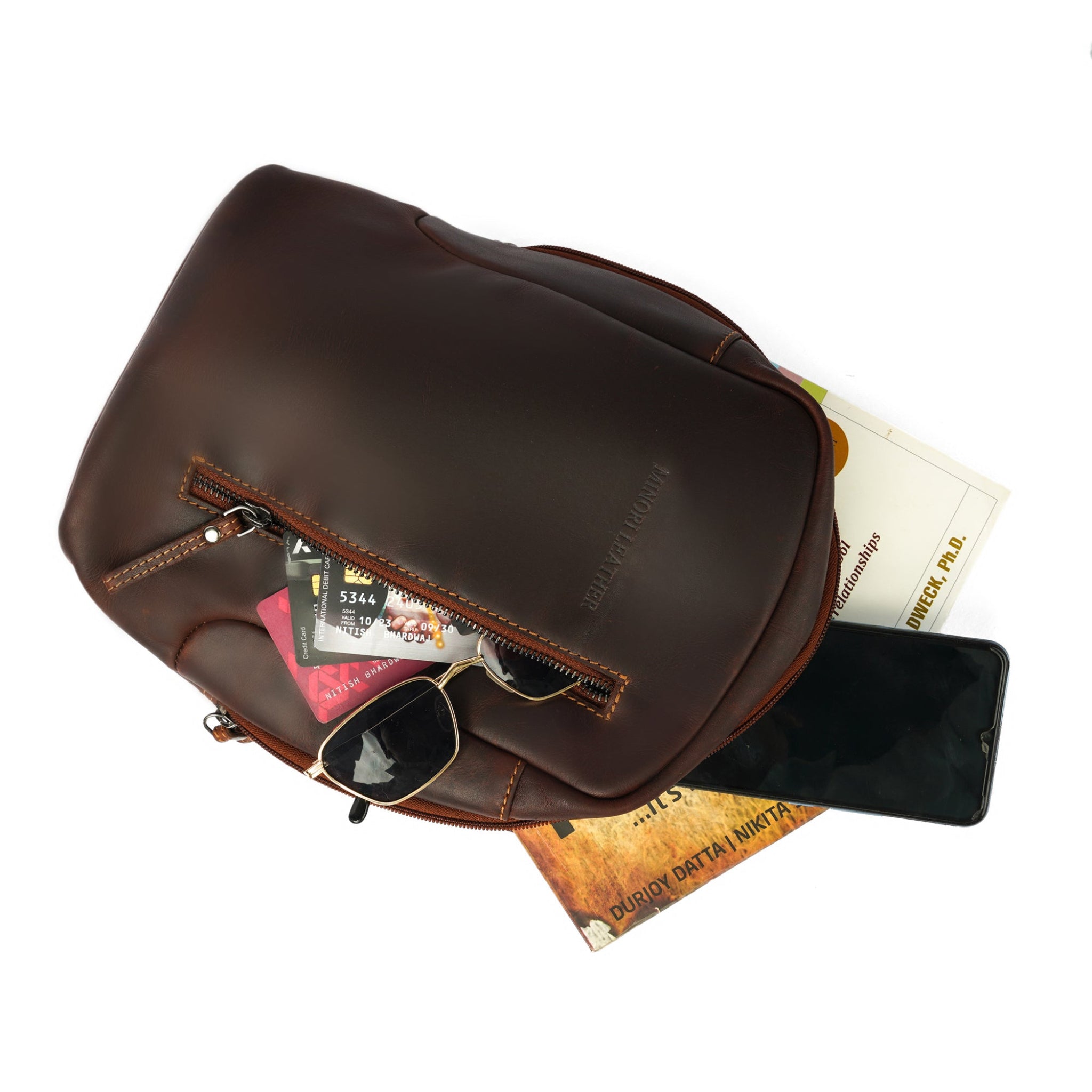 Top-down view of an open brown Minori leather sling bag containing cards, sunglasses, books, and a phone.